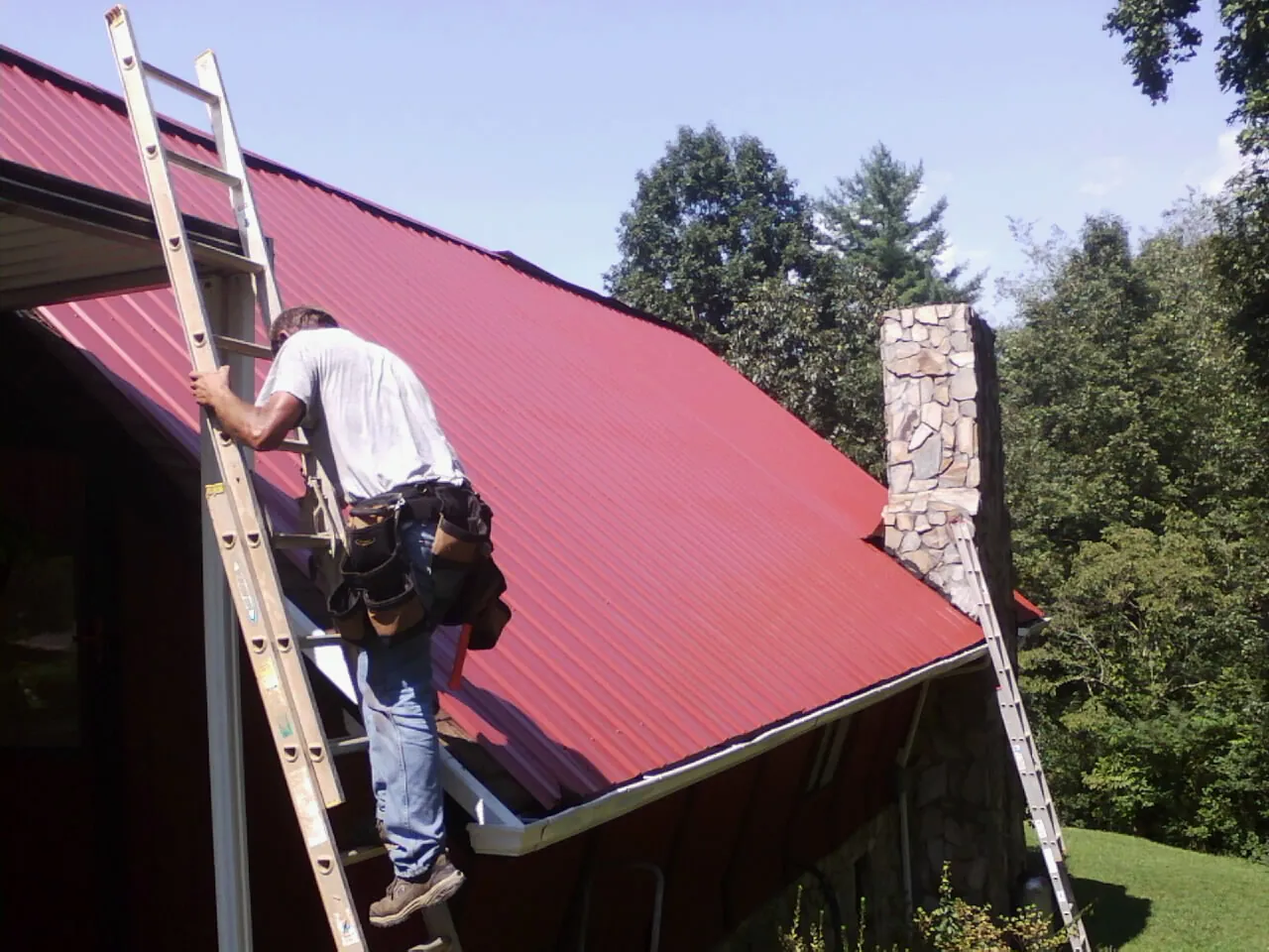 Roofer on a ladder beside a red metal roof and stone chimney — Soffit & Fascia Repair by Roofing Brothers in Jenkinsville